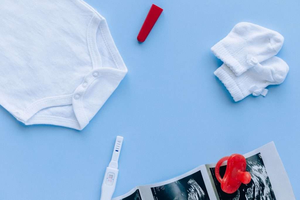 Flat lay of newborn baby essentials including a white bodysuit, socks, pregnancy scan and dummy on a blue background