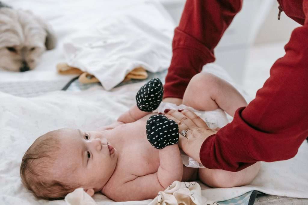 Crop anonymous mother putting on diaper on cute infant baby lying on couch with dog in light room at home