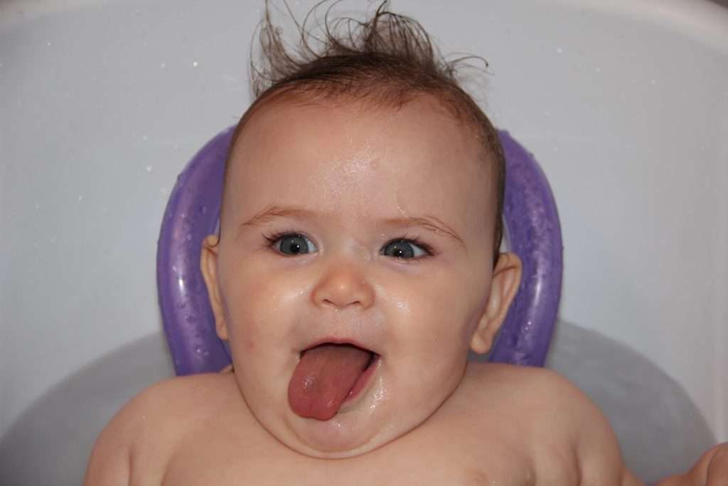 Baby sitting in an upright bath seat with purple ring support, tongue out and happy in the bathtub