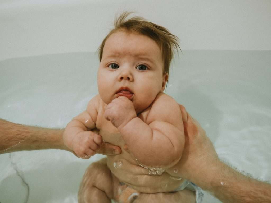 Adult hands supporting an infant in a full-size bathtub, demonstrating constant hands-on supervision during bath time
