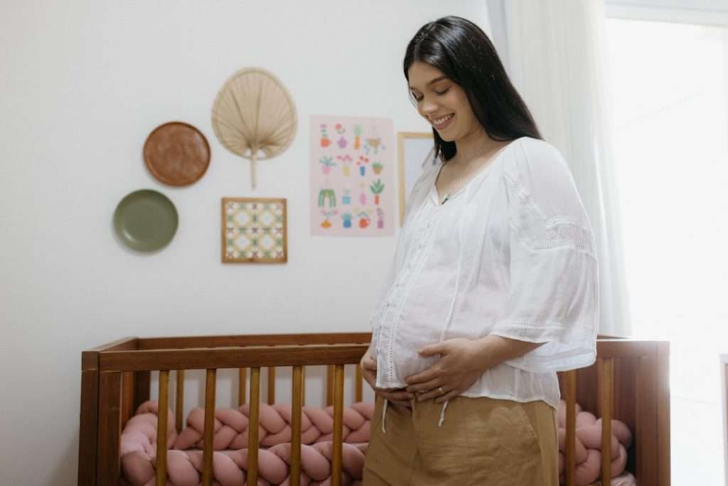 Smiling pregnant mum standing next to a wooden baby cot in a prepared nursery