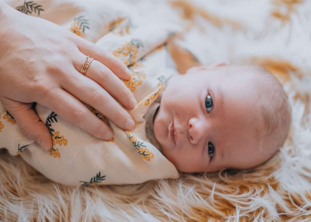 Newborn baby lying on a soft textured blanket wrapped in a muslin swaddle with a parent's hand resting gently on their chest