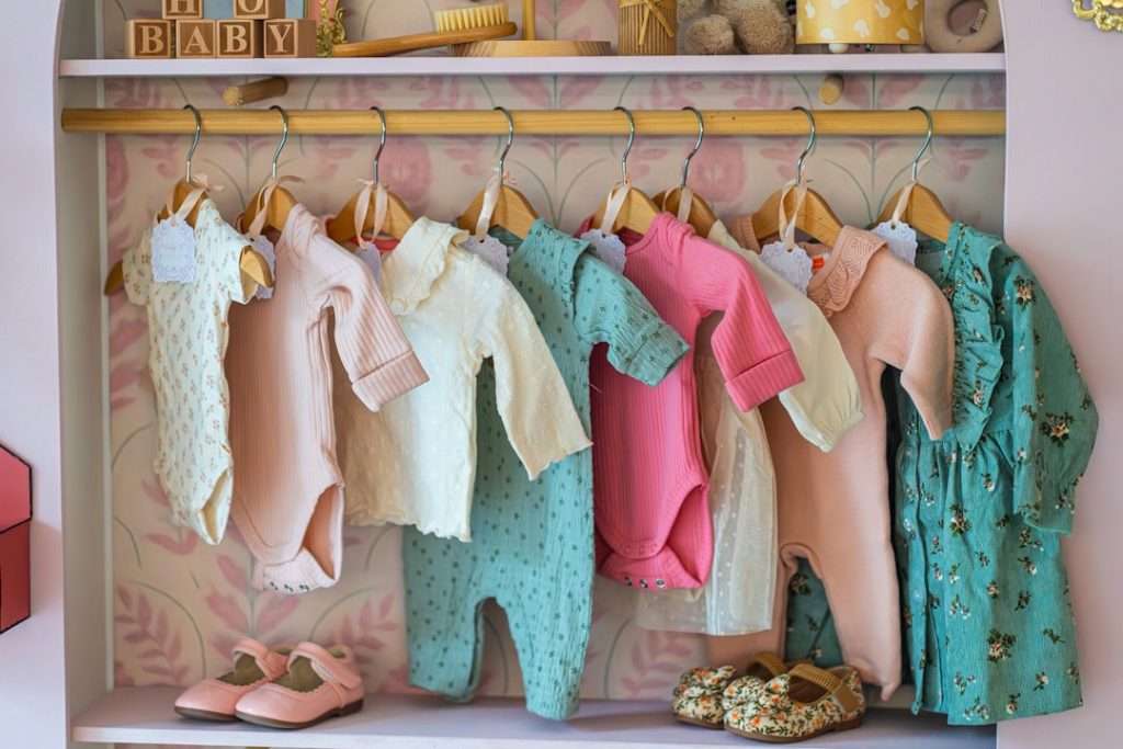 Baby bodysuits and sleepsuits in soft colours hanging neatly on a small nursery rack with tiny shoes below